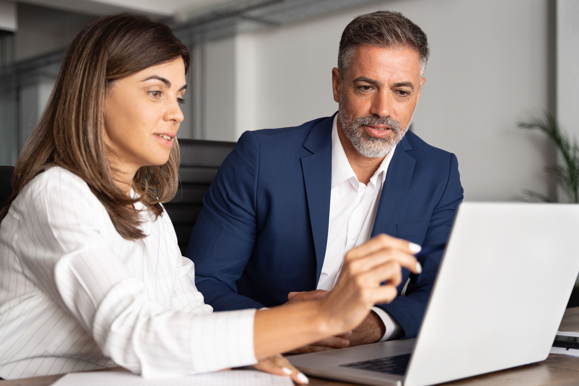 Team of diverse partners sitting at table mature Latin business man and European business woman discussing project on tablet in office. Two colleagues of professional business people working together.